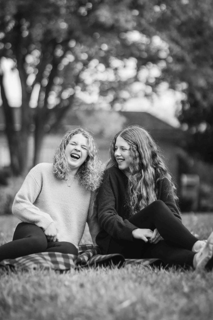 Two young women laughing together while sitting on grass in a park, captured in a family portrait in black and white.