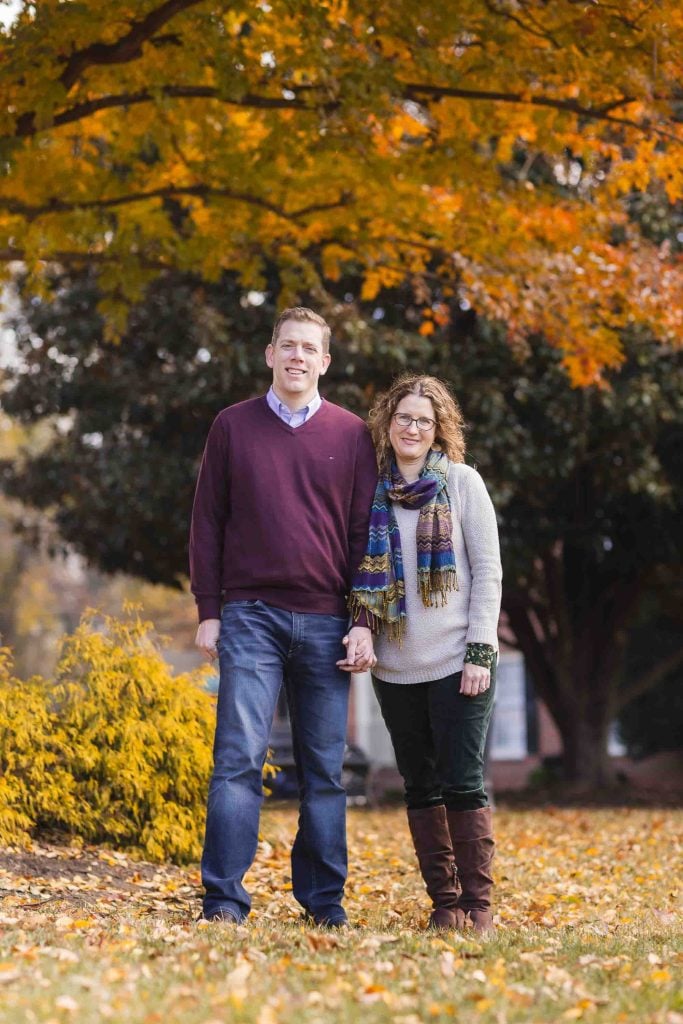 A family portrait of a man and woman standing together in the Kentlands Mansion park with colorful autumn foliage in the background.