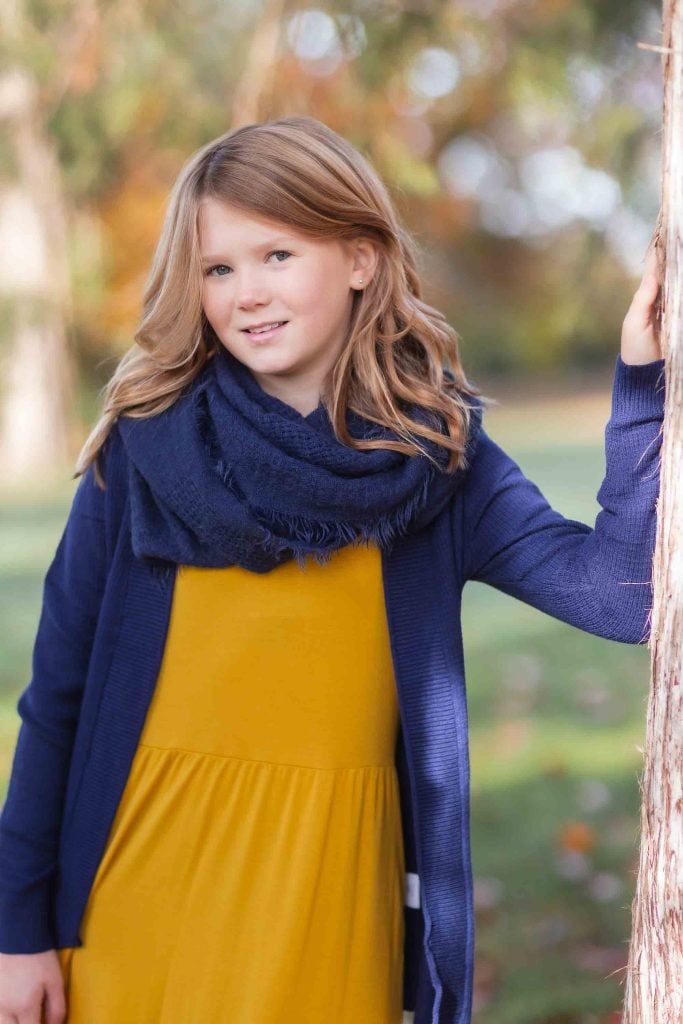 A young girl with blonde hair wearing a yellow dress and blue jacket stands leaning on a tree in a park, perfect for family portraits.