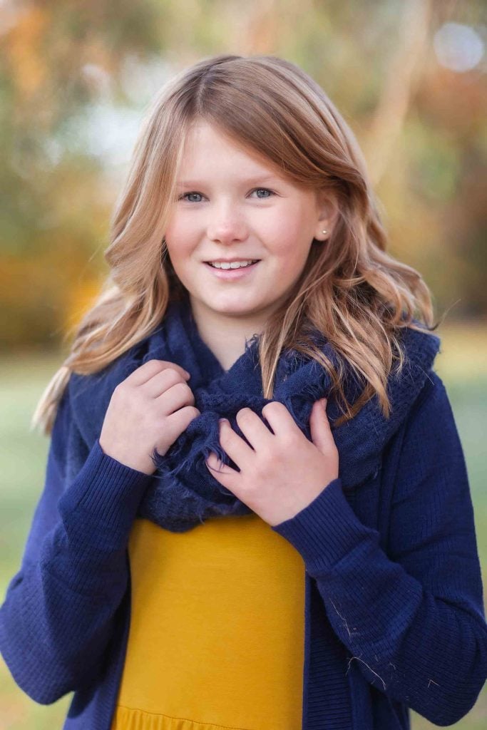 A young girl with long blonde hair, wearing a yellow dress and blue cardigan, smiling in an autumnal park setting for family portraits.