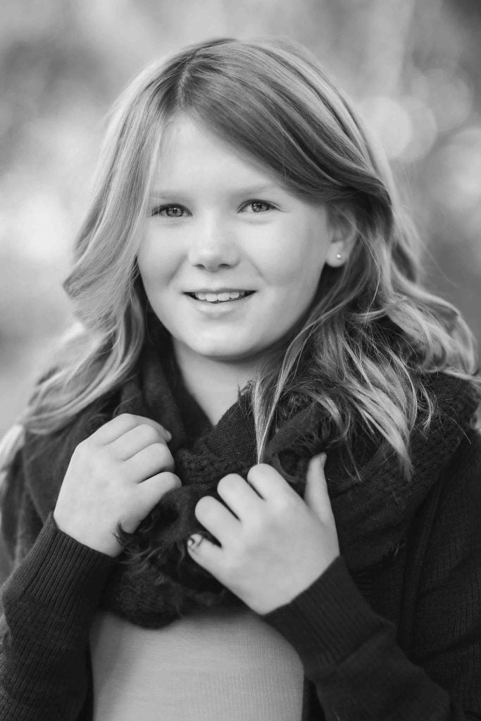 A young girl with blonde hair smiling at the camera, wearing a dark sweater and scarf, with a softly blurred background in black and white. This image is perfect for Family Portraits.