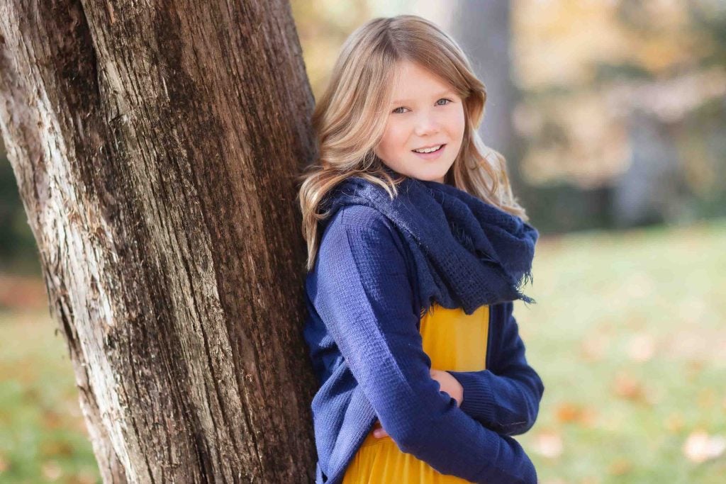 Young girl in a blue sweater and yellow dress smiling near a tree in a park during autumn, perfect for family portraits.