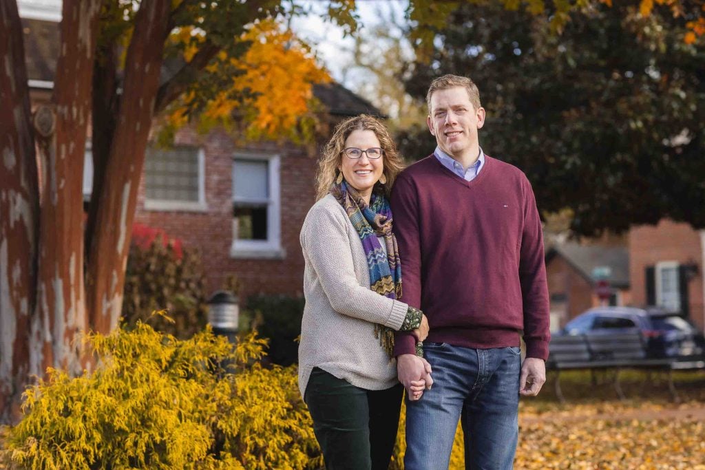 A middleaged couple, smiling and holding hands, stands in front of autumn foliage in a suburban area for their family portraits.