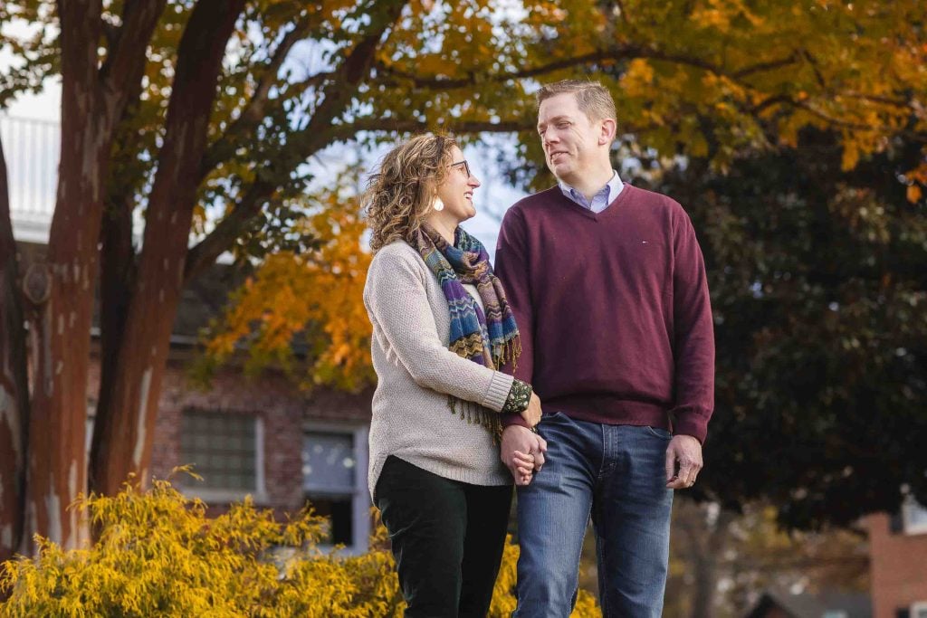 A family enjoying a portrait session in a park with autumn leaves, a couple holding hands and smiling at each other.