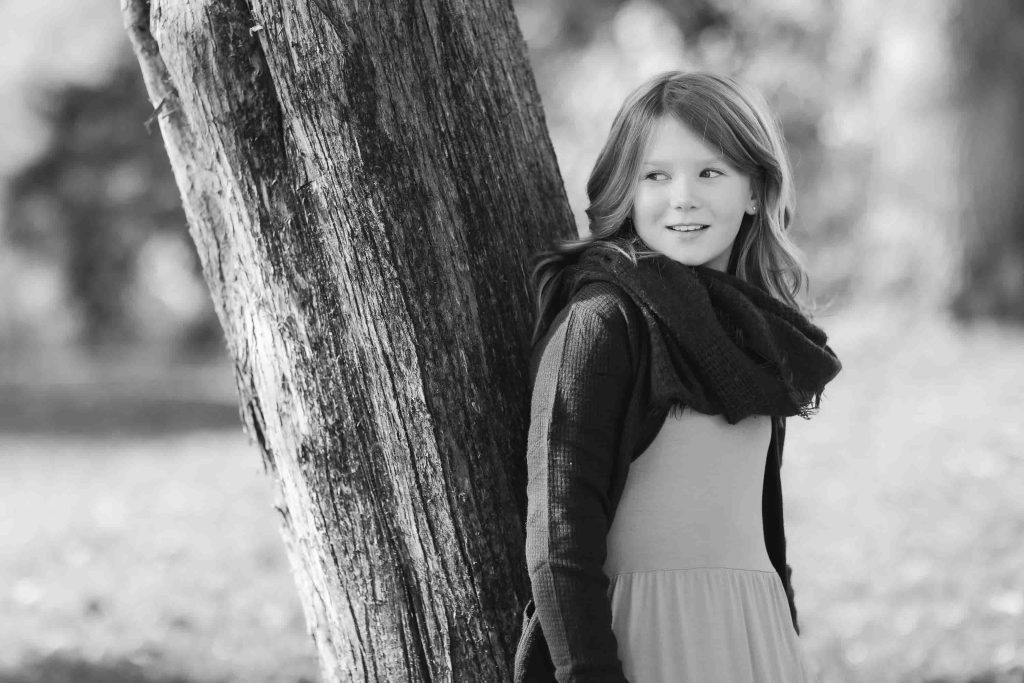 A young girl leaning against a tree in a park, wearing a scarf and a dress, smiling gently in a Family Portraits black and white photo.