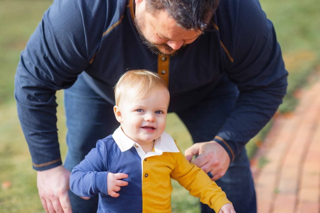 A man crouches down beside a smiling toddler in a yellow and blue outfit, gently supporting the child in a grassy outdoor area for family portraits.