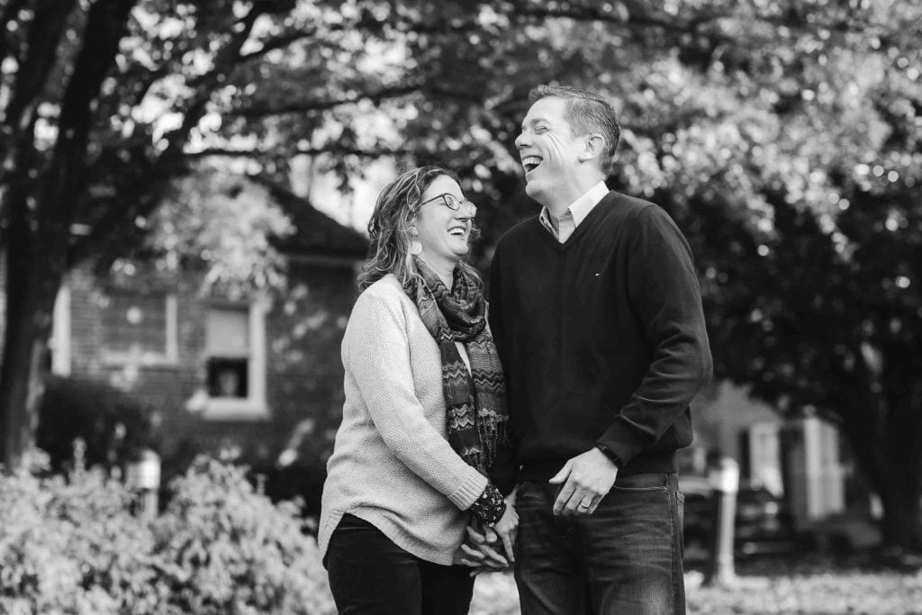 A joyful middleaged couple laughing together in a park, holding hands with trees in the background, in black and white, captured during a family portrait session at Kentlands Mansion.