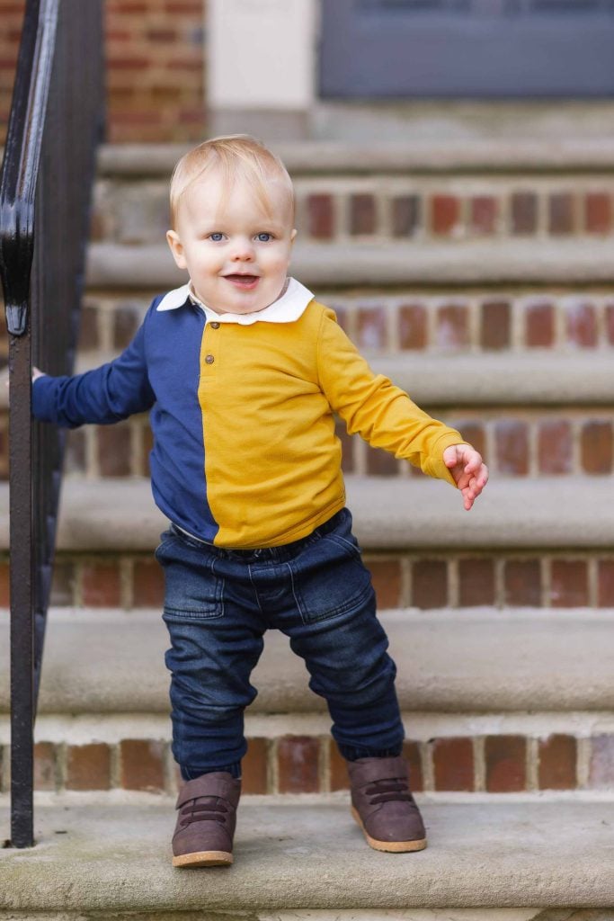 Toddler in a colorblocked shirt and jeans standing on steps, holding a railing, with a cheerful expression for family portraits.