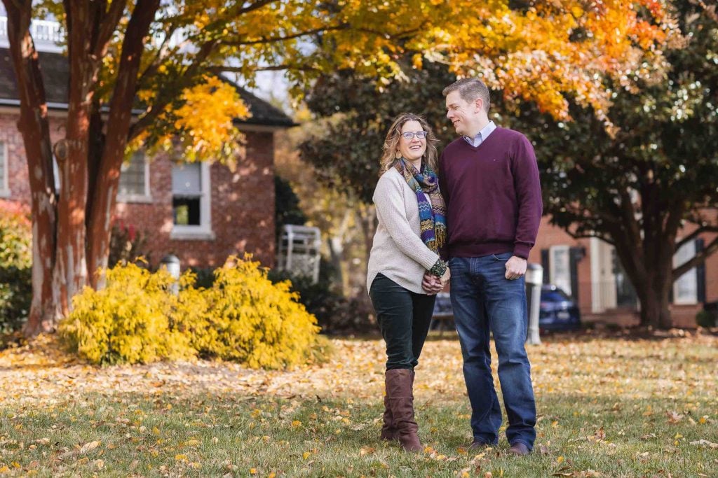 A family portrait of a couple holding hands and smiling at each other in a park with colorful autumn leaves in the background.