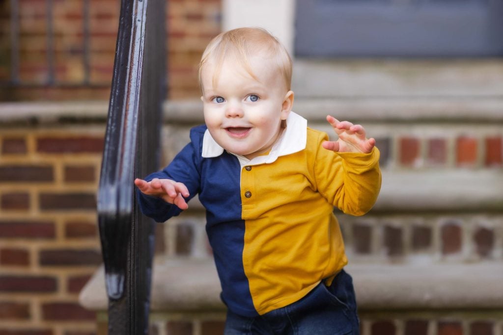 A toddler with blond hair smiles and reaches out, wearing a yellow and blue outfit, standing by a metal railing with a brick background in this charming family portrait.