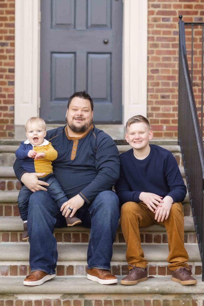 A man sits on steps holding a baby, with a young boy beside them, all smiling in front of a house with a gray door, posing for family portraits.