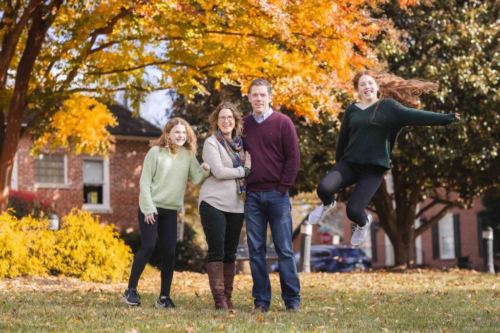 A family of four smiling in an autumnal setting with one member playfully jumping, surrounded by trees with fall colors and a house in the background. This scene is perfect for Family Portraits.