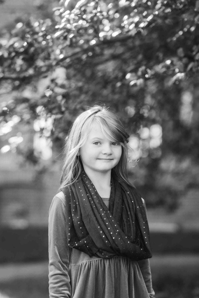 Young girl smiling in a park with trees in the background, dressed in a dress and scarf, depicted in black and white, perfect for family portraits.