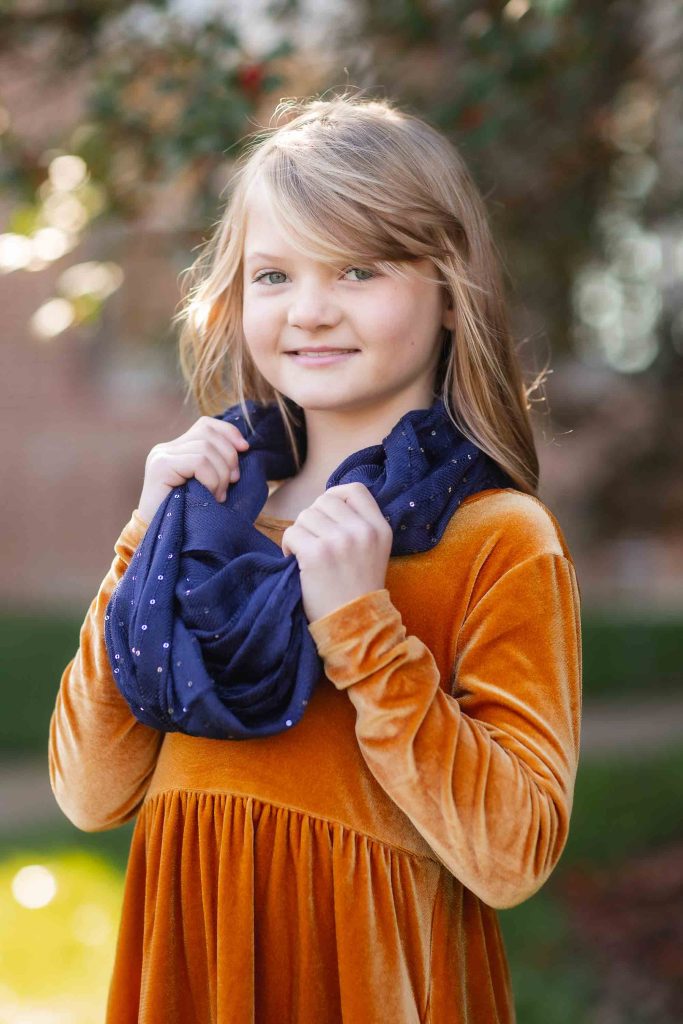 Young girl with blonde hair smiling at the camera, wearing an orange dress and a blue scarf, standing outdoors with blurred greenery in the background, perfect for family portraits.