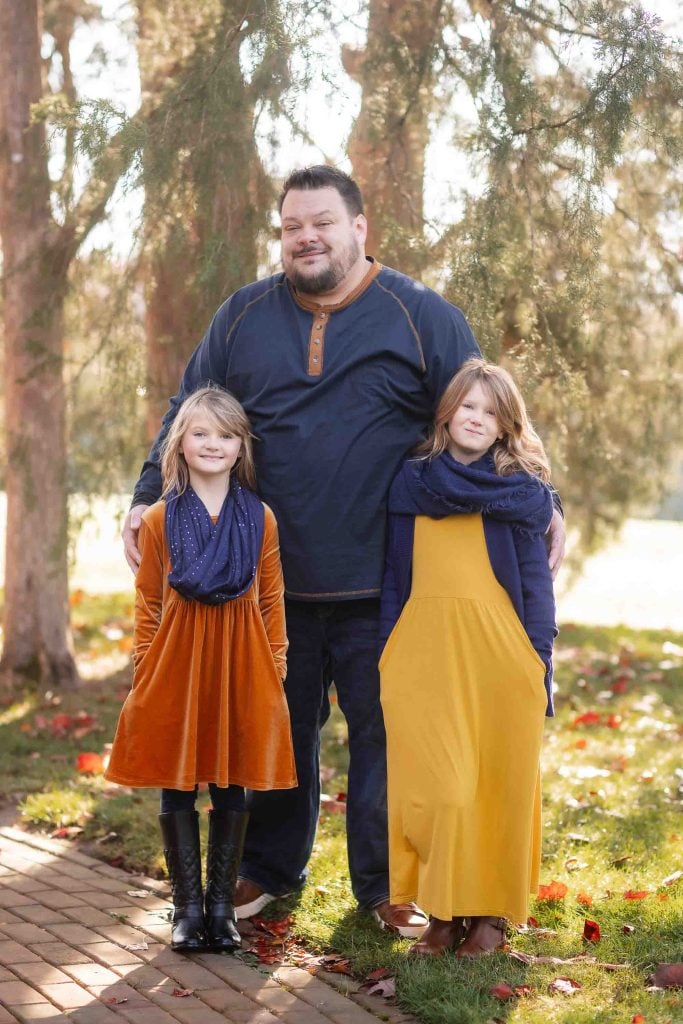 A father stands with his two daughters on a treelined path, all three smiling and dressed in autumn colors for their family portraits.