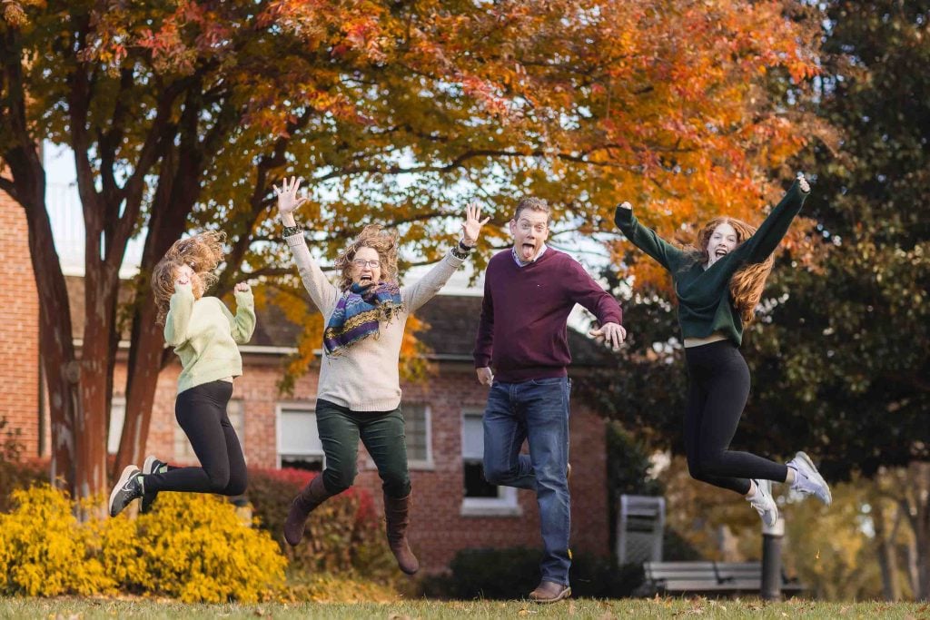 Family portraits of four people joyfully jumping in the air with autumn leaves and trees in the background.