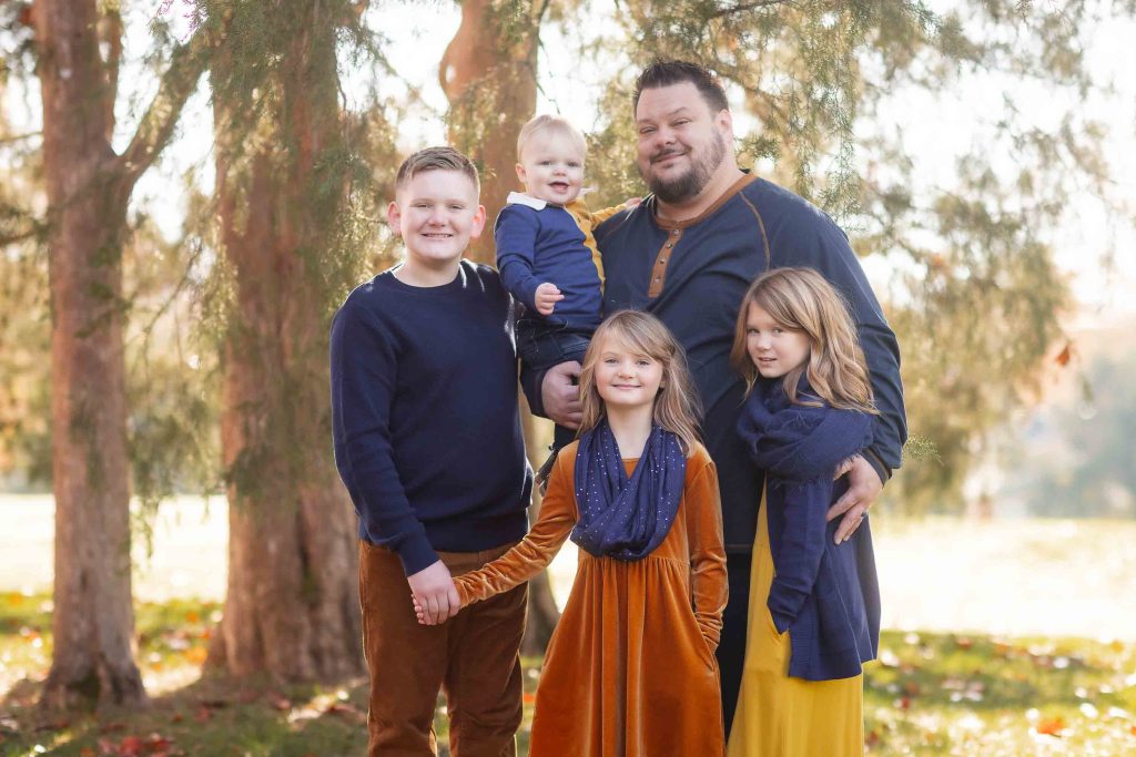 A family of five posing for family portraits outdoors, with two adults and three children, standing among trees in a sunny park.