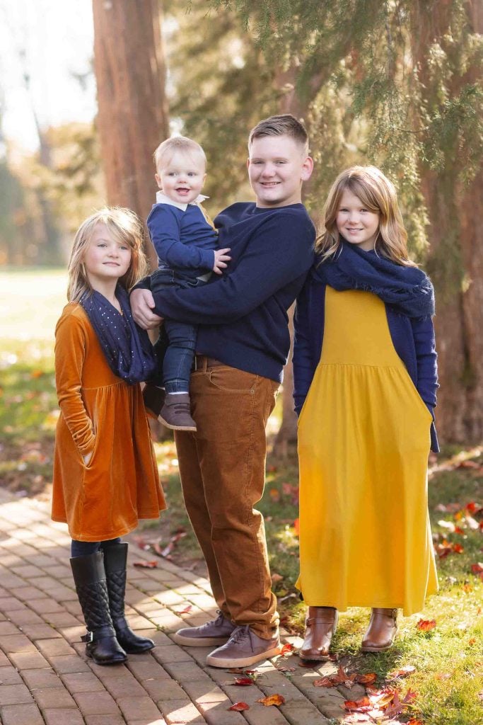 A Family Portraits of four, including two young girls and a baby boy held by their father, posing together in a park with tall trees in the background.