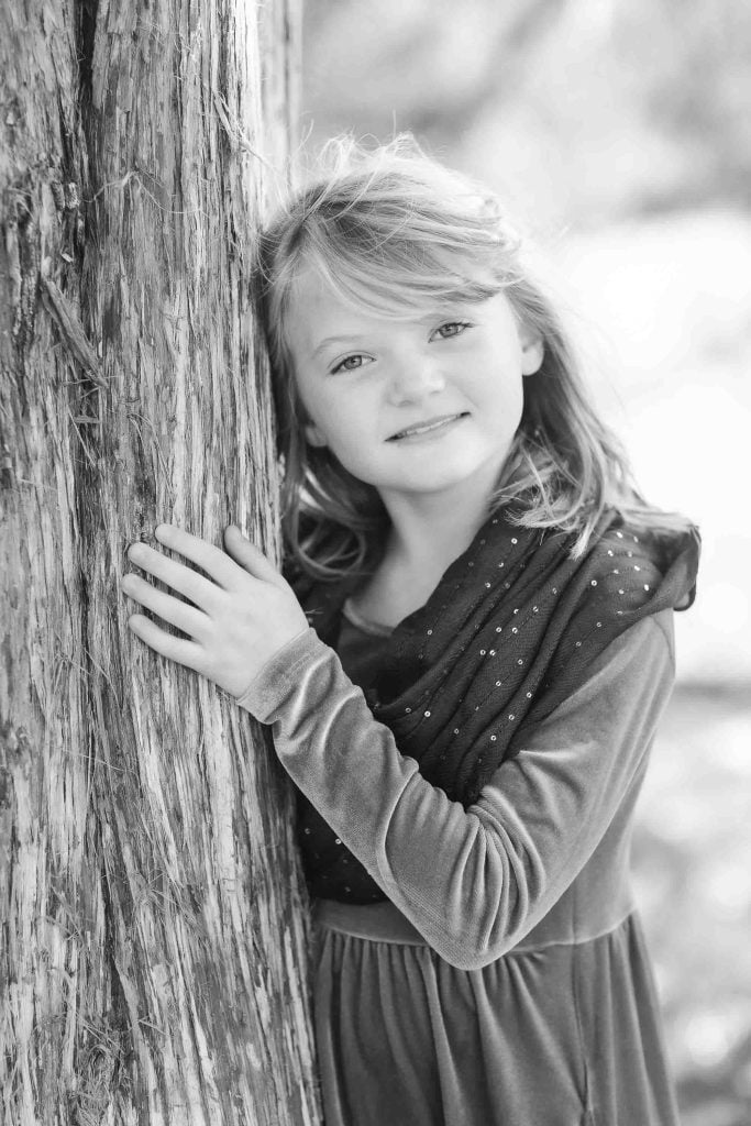 Young girl leaning against a tree, looking at the camera with a gentle smile, in a black and white family portrait photograph.