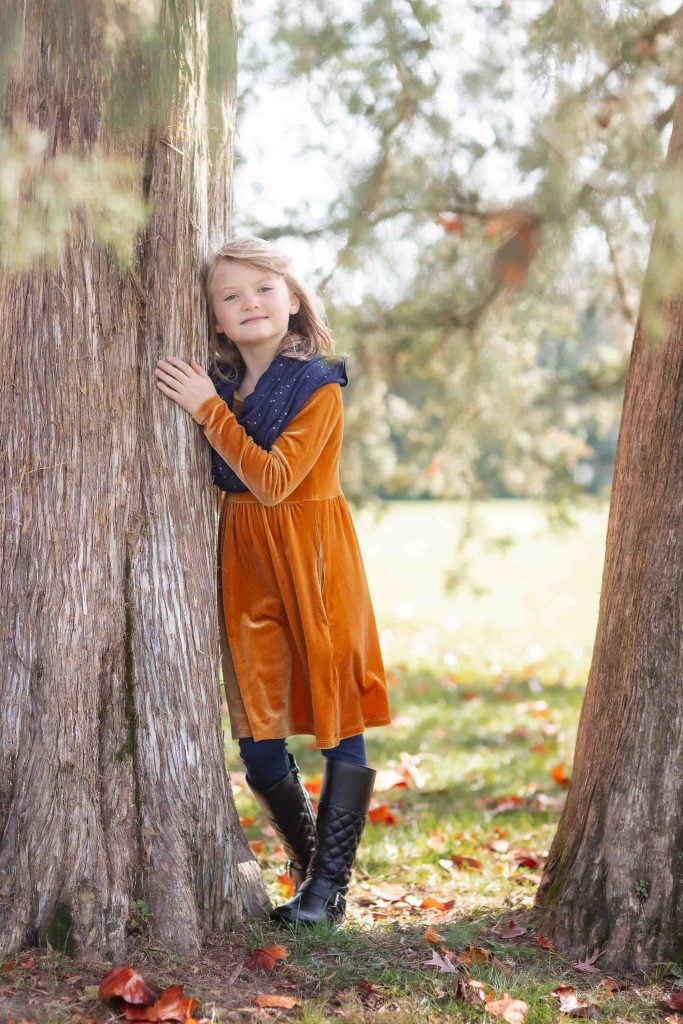 A young girl in an orange dress and black boots smiling and peeking from behind a tree in a sunlit park, perfect for family portraits.