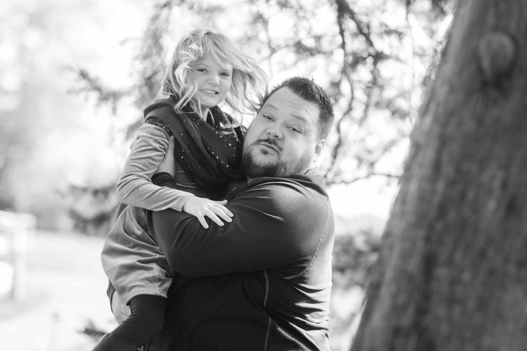 A man carrying a young girl on his back in a sunny park, both smiling, in a black and white family portrait photo.