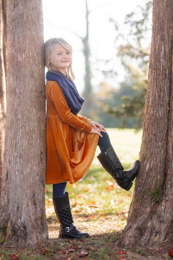 Young girl in an orange dress and black boots sitting between two trees in a sunny park, smiling gently at the camera for family portraits.