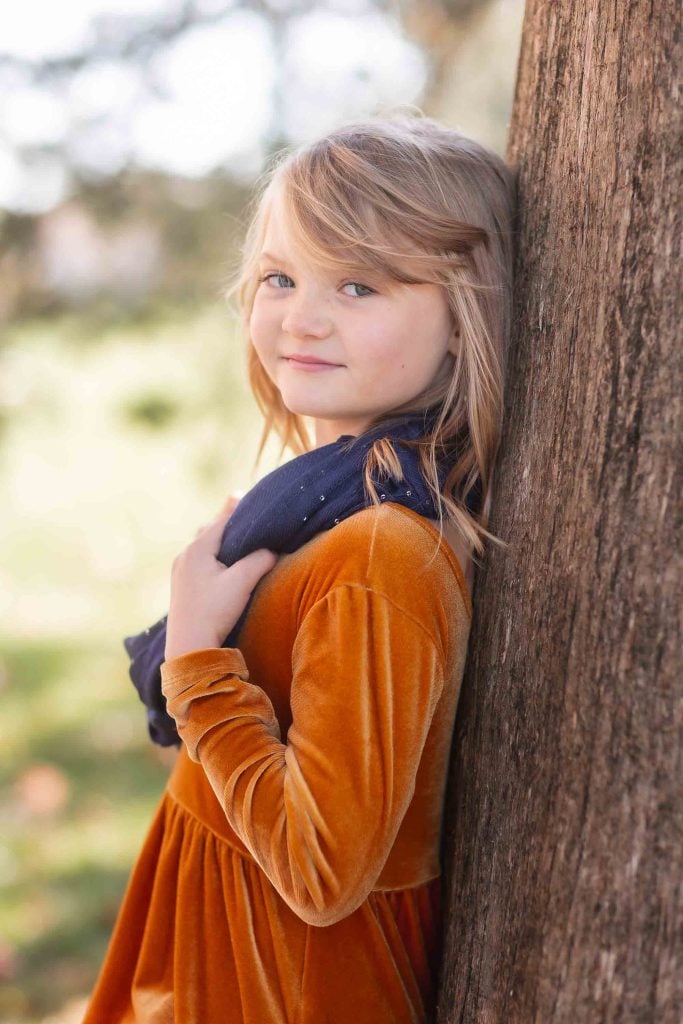 A young girl with blond hair, wearing an orange dress and navy scarf, leaning against a tree and looking over her shoulder in a family portrait.