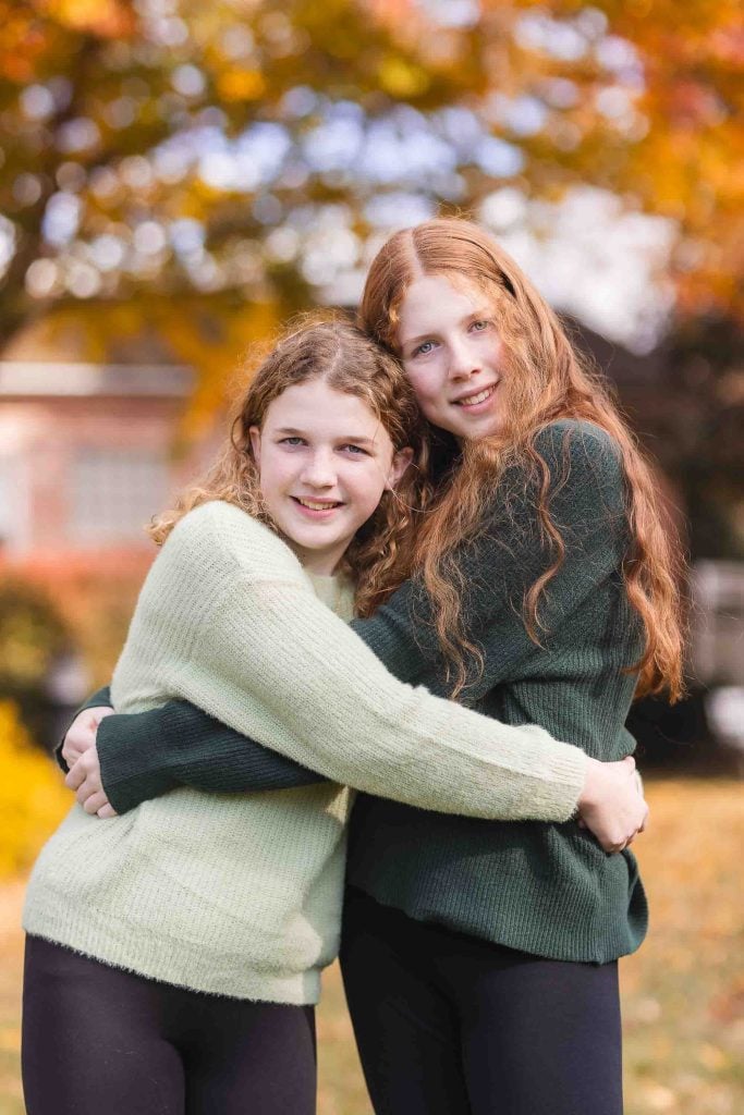 Two young girls with curly hair, one brunette and one redhead, smiling and hugging in a park during autumn, perfect for family portraits.