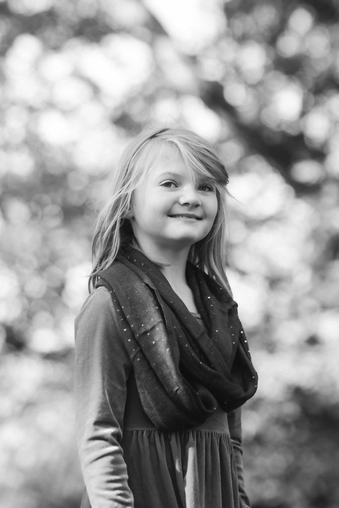 Young girl with blond hair smiling in a park, wearing a dress and scarf, with blurred trees in the background. Family Portraits.