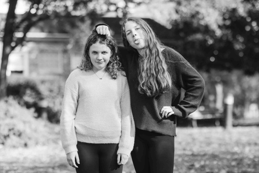 Two young girls posing outdoors for a family portrait, one playfully resting her fist on the other's head, both smiling in a park setting.