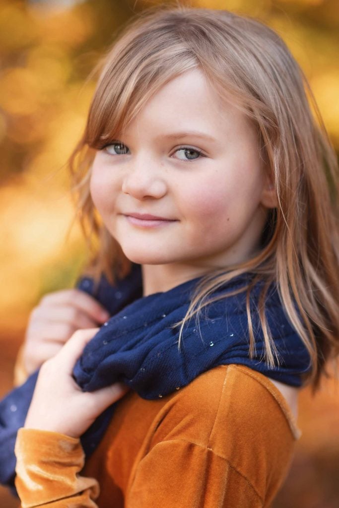 A young girl with light brown hair, wearing an orange dress and a blue scarf, smiling gently against an autumn leaf background for family portraits.