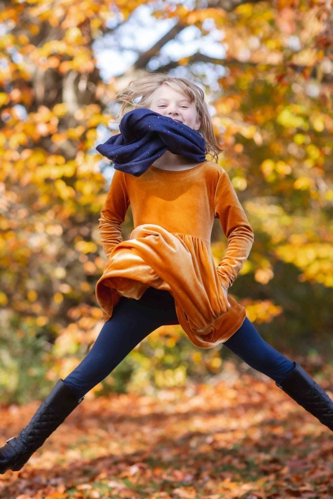 Young girl in orange dress and black leggings joyfully jumping with a blue scarf in an autumn park with golden leaves for Family Portraits.