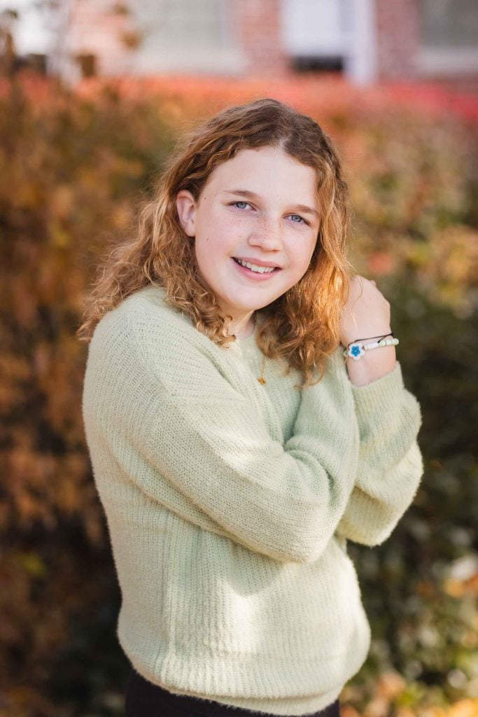 Young girl with curly hair smiling at the camera in a family portrait, wearing a green sweater, standing outdoors.