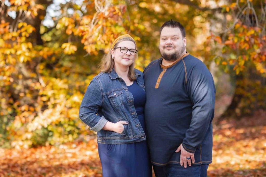 A couple standing closely together in an autumn park for a family portrait, surrounded by trees with orange leaves, smiling gently at the camera.