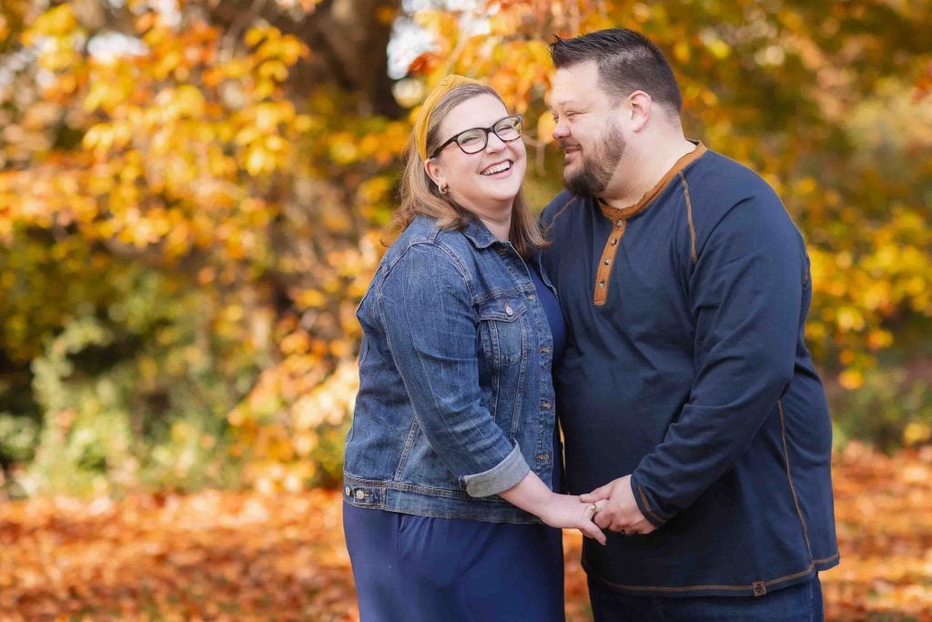 A happy couple holding hands and smiling at each other in a park with autumn leaves in the background, perfect for family portraits.