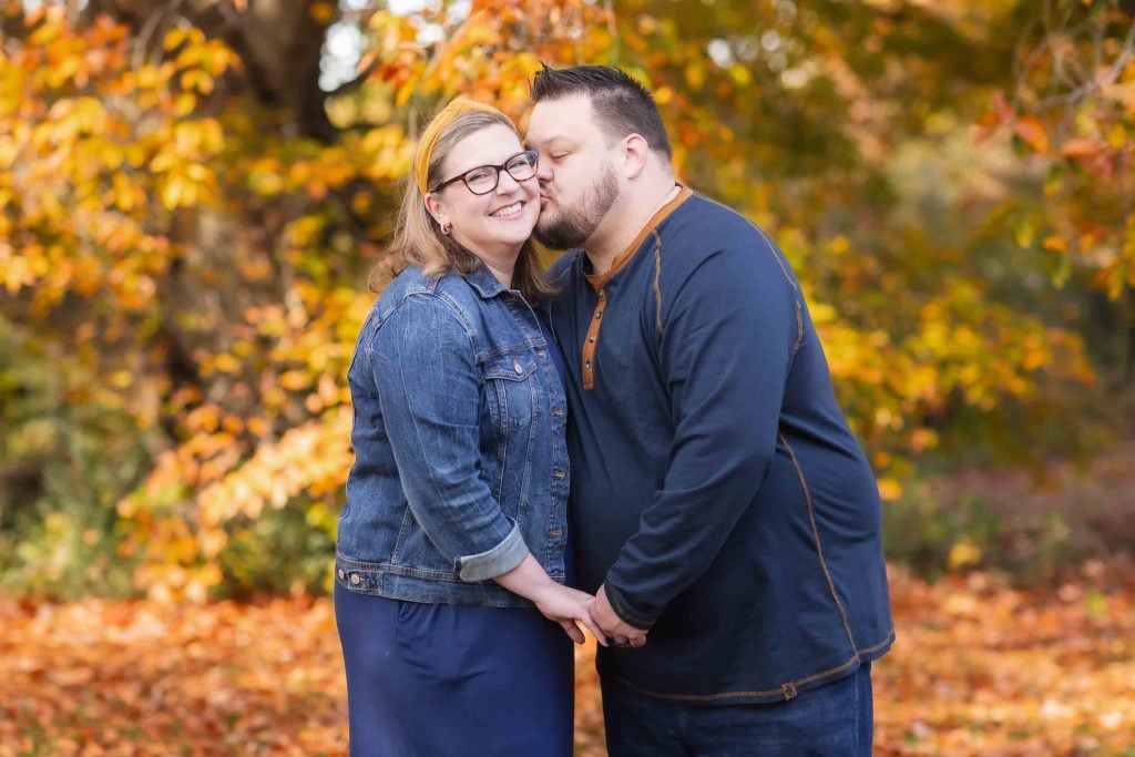 A couple embracing and smiling in an autumnal park, with the man kissing the woman's forehead, perfect for Family Portraits.