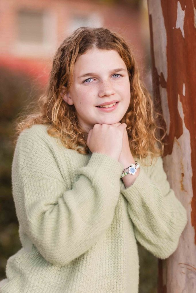 A young girl with curly hair smiles while leaning on a tree, wearing a green sweater and a watch, perfect for family portraits.