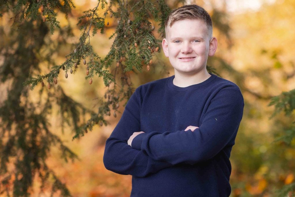 A young boy wearing a blue sweater smiles at the camera with his arms crossed, standing in front of autumn trees with golden leaves, perfect for family portraits.