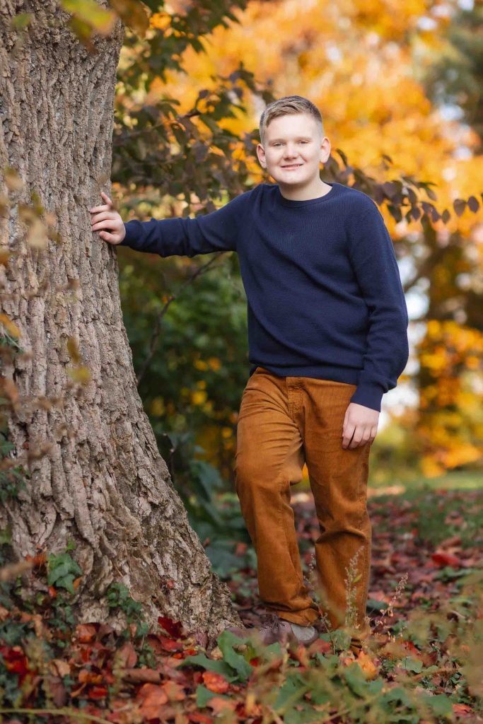 A young boy in a navy sweater and brown pants stands by a tree surrounded by autumn leaves, smiling at the camera for family portraits.