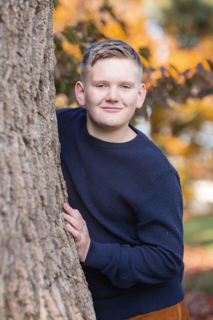 A young boy in a navy sweater smiling next to a tree with autumn leaves, perfect for family portraits.