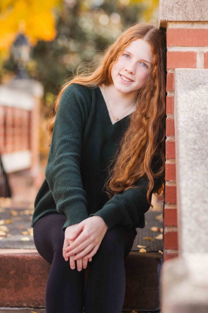 Young woman with long red hair sitting on brick steps, wearing a green sweater, smiling at the camera in an autumn setting for Family Portraits.