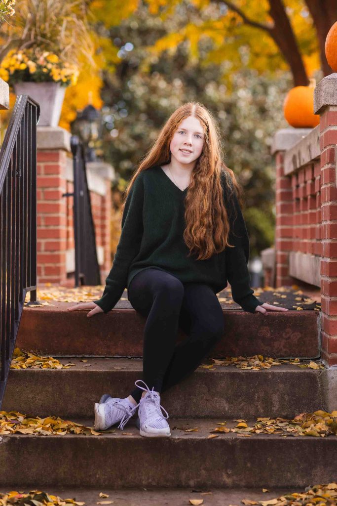 A young woman with long red hair sitting on steps surrounded by autumn leaves and pumpkins, wearing a dark green sweater and black leggings, perfect for family portraits.