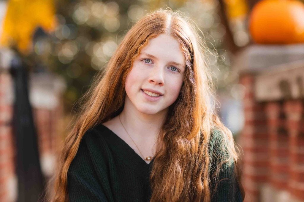 Young woman with red hair smiling outdoors in front of a blurred autumn background for family portraits.