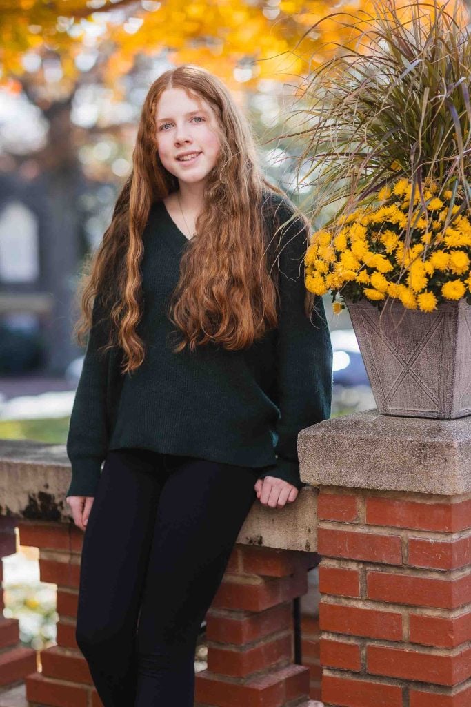 A young woman with long curly hair sitting on a brick wall at Kentlands Mansion, smiling, with a pot of yellow flowers next to her.