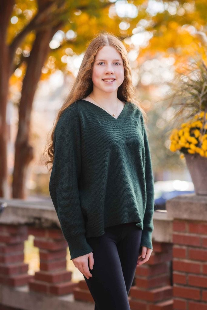 A young girl with long curly hair, wearing a green sweater, posing in front of autumncolored trees for family portraits.