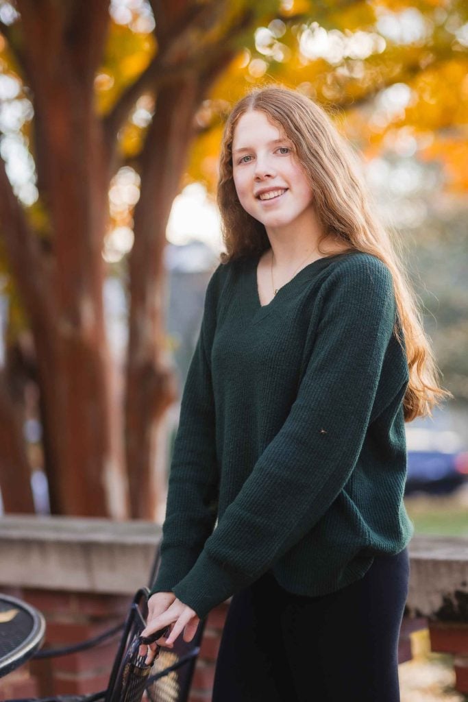 Young woman with long blonde hair wearing a green sweater, smiling in a park during autumn, standing beside a bicycle for Family Portraits.