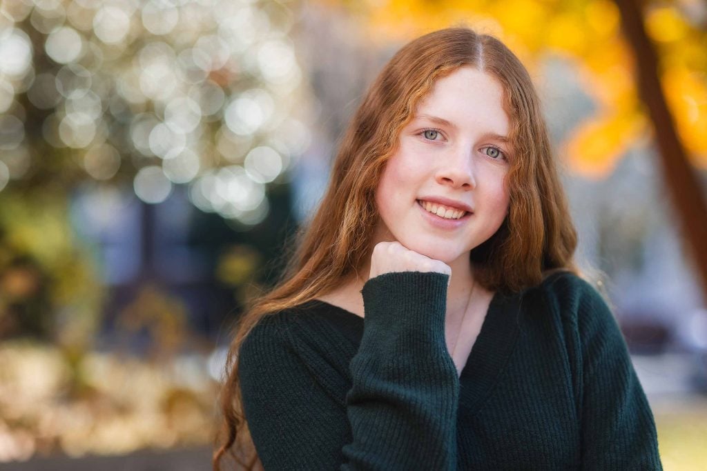 A young woman with long red hair smiling, resting her chin on her hand, with golden autumn leaves blurred in the background for a Family Portrait.