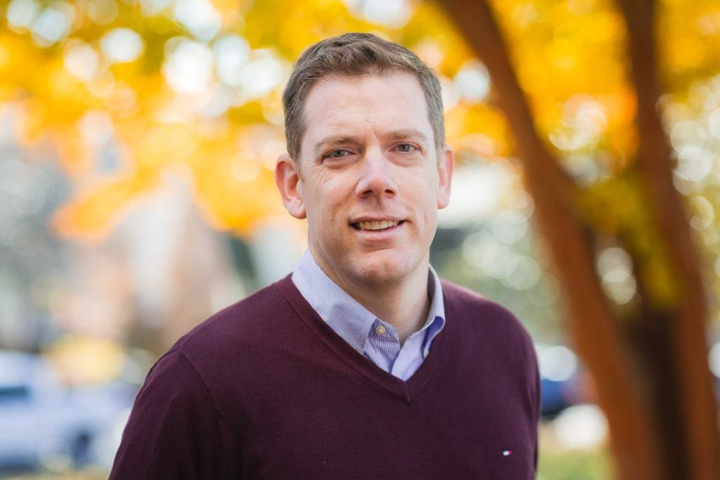 A smiling man in a maroon sweater and checkered shirt, standing outdoors with autumn trees in the background, perfect for family portraits.