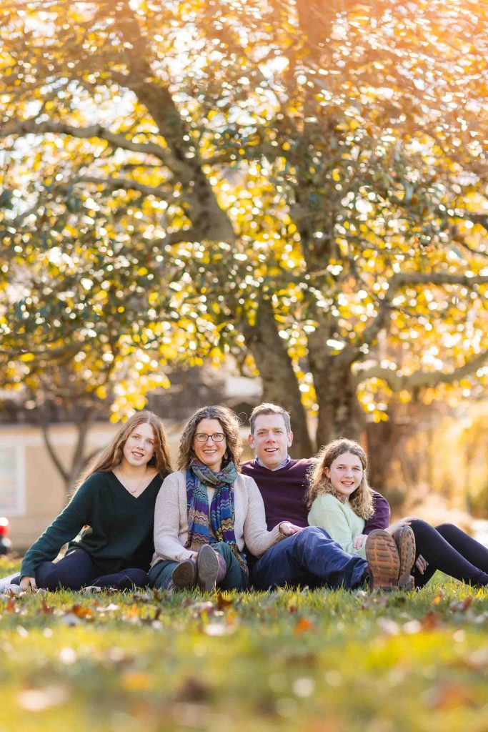 A family portrait of four sitting side by side on grass with a backdrop of sunlit autumn trees, smiling at the camera.