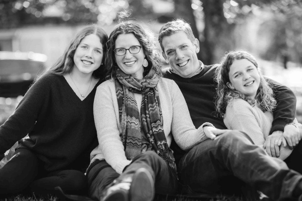 A family of four, including two adults and two teens, sitting together on grass and smiling at the camera in a black and white Family Portrait.